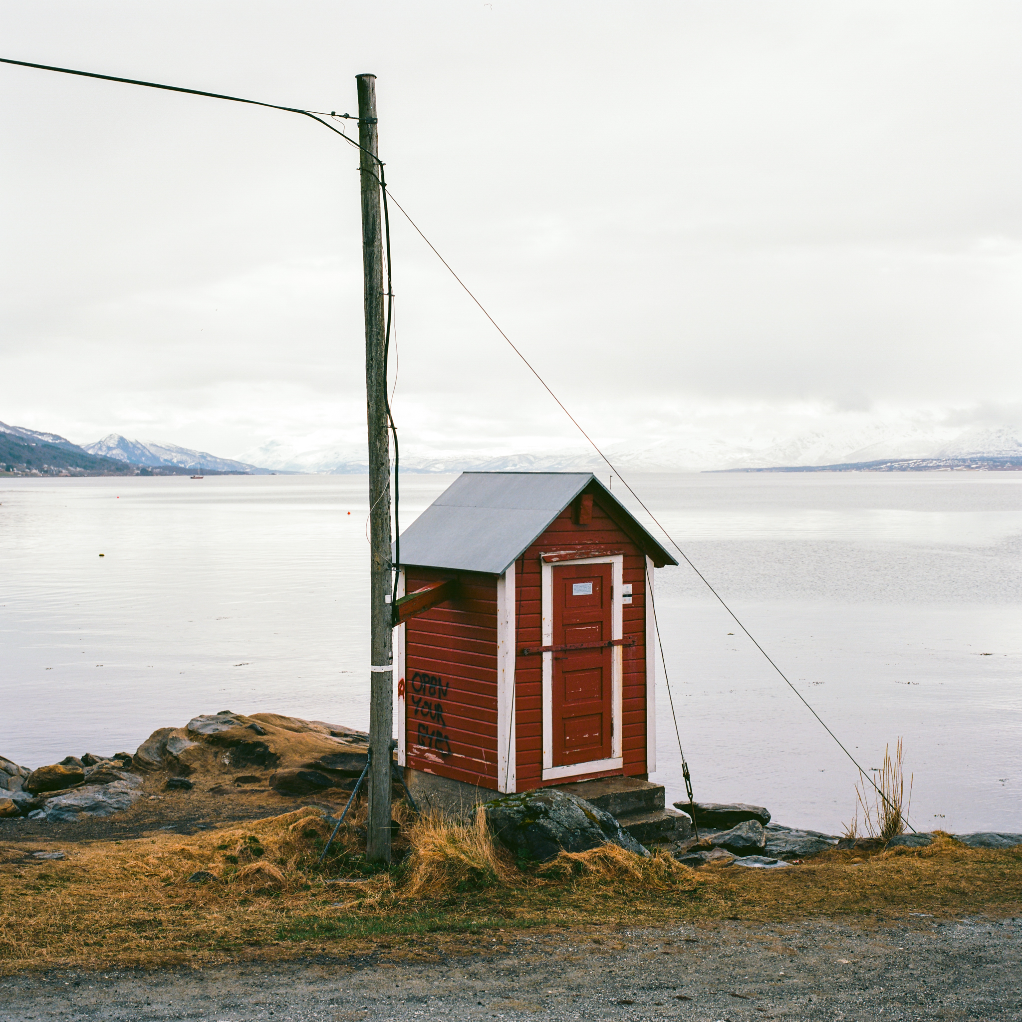 A tiny wooden shack spray painted open your eyes, overlooking the water outside Tromsø with the mountains in the distance