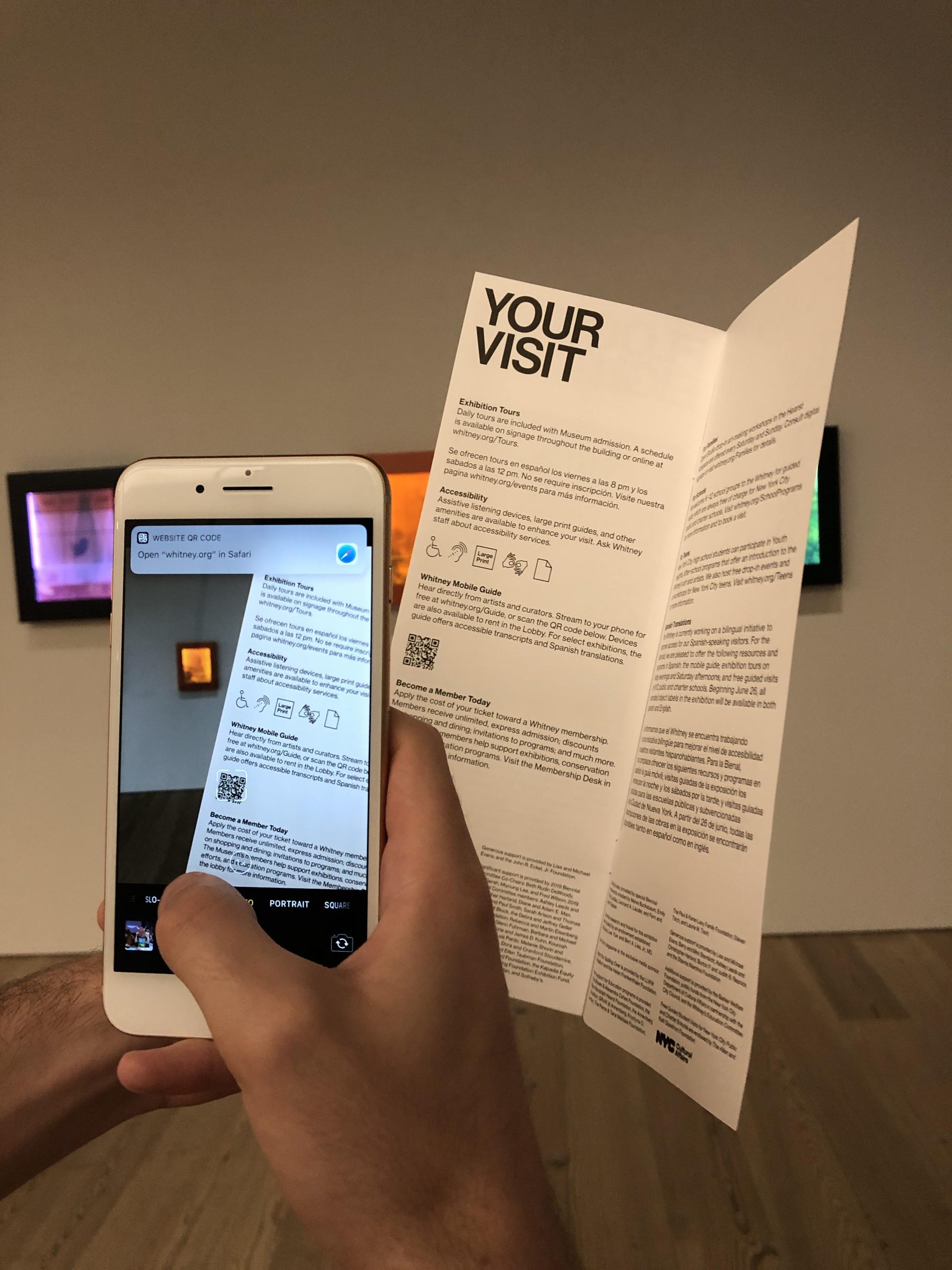 An iPhone with the camera app open held up to scan a QR code on a paper museum guide.